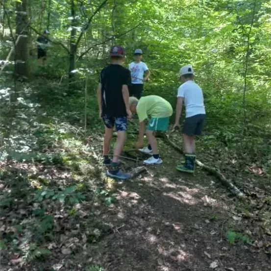 Fünf Kinder wandern auf einem schmalen Pfad in einem dichten Wald. Ein Kind beugt sich hinunter, um etwas am Boden zu untersuchen.