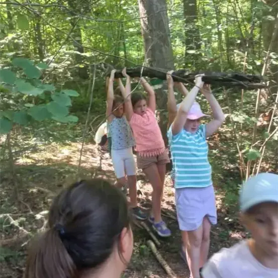 Vier Kinder klettern in einem Wald auf einen Ast. Sie halten sich an einem Stock fest und scheinen für ein Foto zu posieren. Eine erwachsene Frau beobachtet sie von hinten.