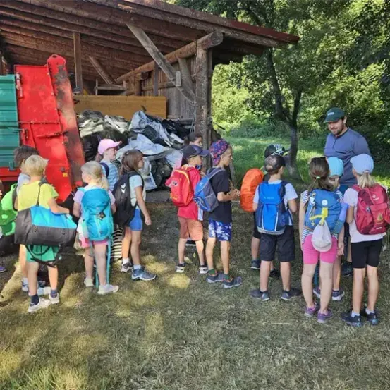 Eine Gruppe von Kindern mit Rucksäcken steht auf einem Feld, während ein Mann mit grünem Hut zu ihnen spricht. Dahinter kippt ein roter Lastwagen Abfall aus.