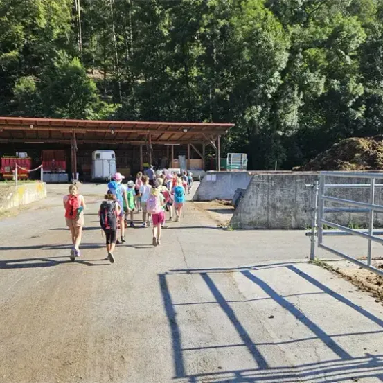 Eine Gruppe von Kindern geht auf einem Betonweg in Richtung einer Scheune mit rotem Dach. Rechts befindet sich ein Metallzaun, in der Nähe ein Haufen Erde. Bäume und ein Hang sind im Hintergrund.