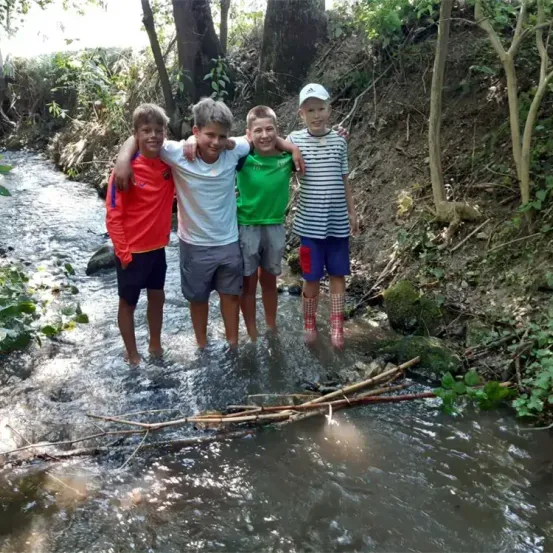 Vier Jungen stehen in einem kleinen Bach und lächeln für ein Foto. Sie tragen verschiedenfarbige Hemden und Shorts.