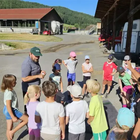 Eine Gruppe von Kindern steht vor einem Mann in einem grünen Hut auf einem Parkplatz mit Gebäuden und Hügeln im Hintergrund.