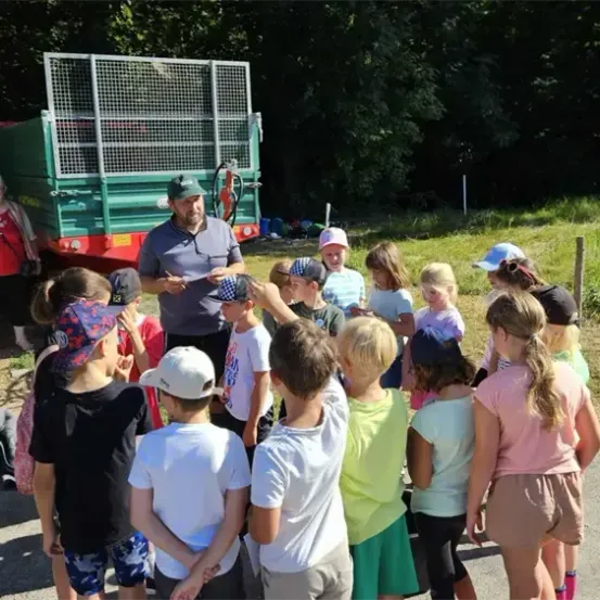 Eine Gruppe von Kindern ist um einen Mann vor einem grünen Anhänger versammelt. Der Mann hält ein Stück Papier in der Hand und spricht zu den Kindern. Eine erwachsene Frau steht in der Nähe des Anhängers.