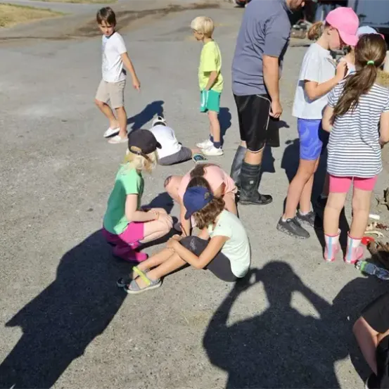 Eine Gruppe von Kindern und Erwachsenen versammelt sich auf einer Straße. Einige sitzen, andere stehen. Sie tragen Freizeitkleidung. Die Schatten der Menschen fallen auf die Straße.
