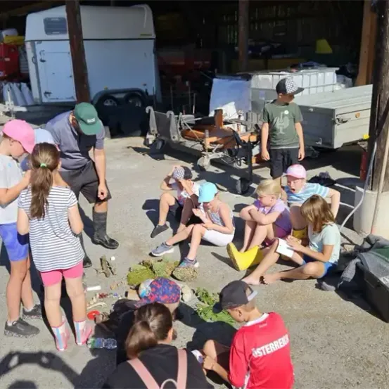 Eine Gruppe von Kindern und Erwachsenen versammelt sich vor einer Scheune. Sie sitzen und stehen auf dem Boden, wobei einige Kinder kleine Pflanzen am Boden betrachten. Ein Mann steht vor ihnen, und es gibt verschiedene Gegenstände in der Umgebung.