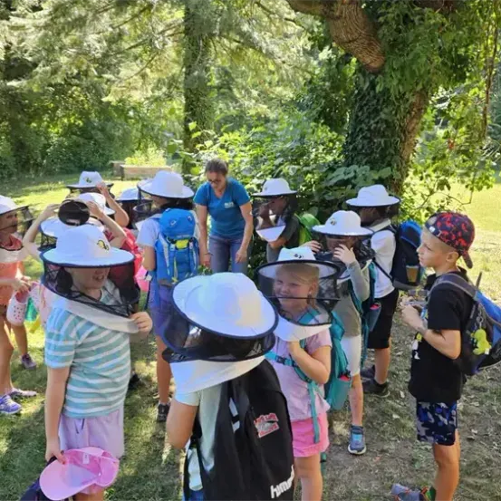 Eine Gruppe von Kindern mit Imkerhüten und Rucksäcken auf einer Grasfläche, mit einer erwachsenen Frau in einem blauen Shirt.