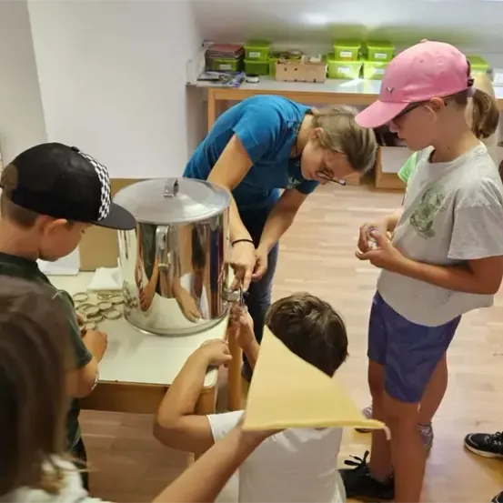 Eine Frau hilft Kindern um einen Metallbehälter herum, wahrscheinlich in einem Klassenzimmer. Sie halten Papierkegel, einige beobachten. Ein Poster ist hinter ihnen.
