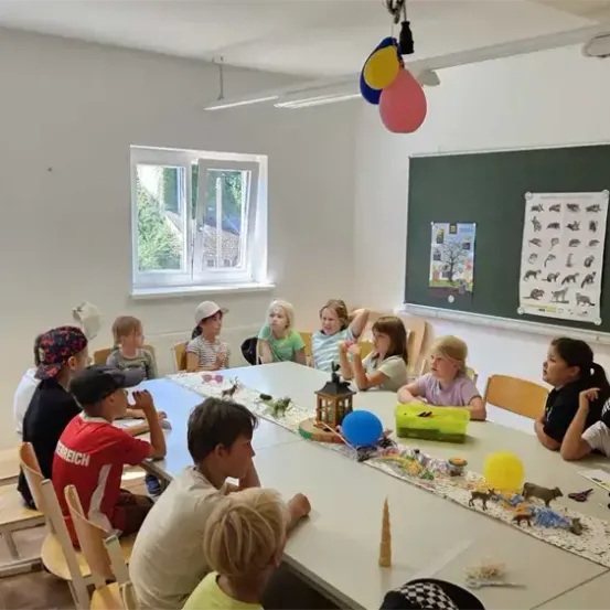 Eine Gruppe von Kindern sitzt um einen langen Tisch in einem Klassenzimmer, einige tragen Mützen. Ein Tafel mit Zeichnungen steht dahinter, und der Raum hat weiße Wände und Fenster.