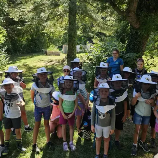 Eine Gruppe von Kindern in Imkeranzügen steht in einem Garten mit einer Frau in der Nähe. Sie tragen Schutzkappen und scheinen etwas über Imkerei zu lernen.