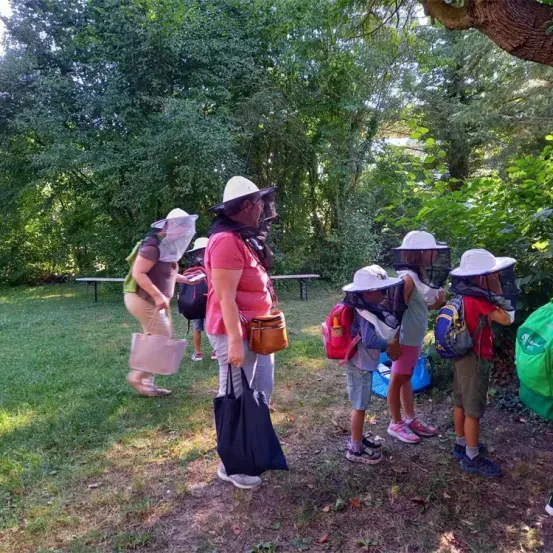 In einem Park versammeln sich mehrere Kinder und Erwachsene, die Imkermützen tragen und Taschen halten. Die Kinder stehen vor einem Busch, während Erwachsene in der Nähe stehen. Bäume und eine Bank sind im Hintergrund zu sehen.