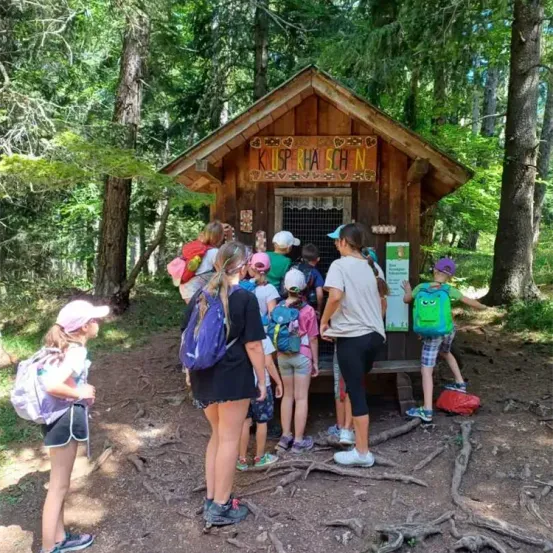 Eine Gruppe von Kindern steht vor einer kleinen Holzhütte mit der Aufschrift 'KILPSPERHA SCHN' im Wald. Sie tragen alle Rucksäcke und einige Hüte.