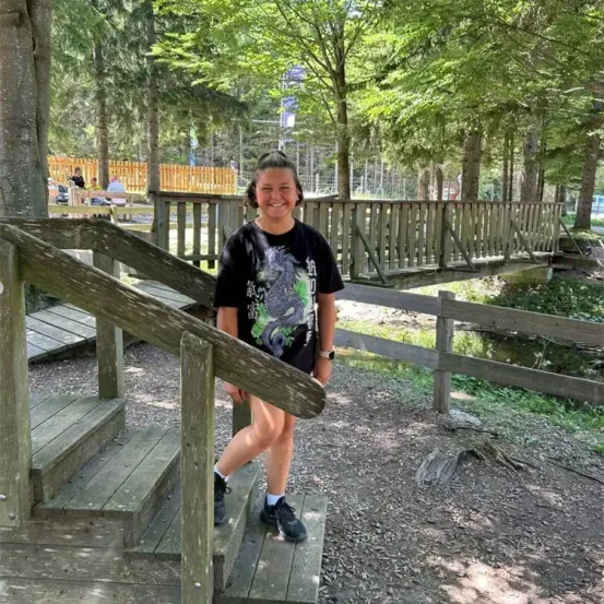 Eine lächelnde Frau in einem schwarzen T-Shirt steht auf einer Holztreppe in der Nähe einer Holzbrücke, mit einem eingezäunten Bereich und Bäumen dahinter.