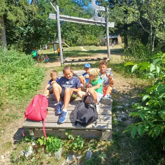 Fünf Kinder sitzen auf einer Holzplattform in einem Waldgebiet. Zwei von ihnen tragen Mützen und sie alle lächeln. In der Nähe sind Rucksäcke. Ein Holzgerüst steht hinter ihnen.