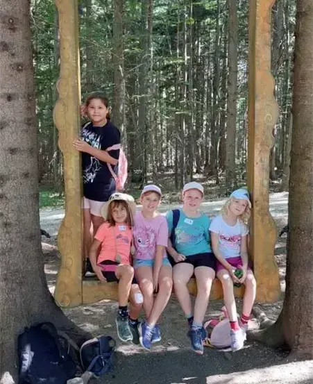 Fünf junge Mädchen sitzen auf einer Holzbank im Wald mit Rucksäcken in der Nähe. Sie posieren für ein Foto.