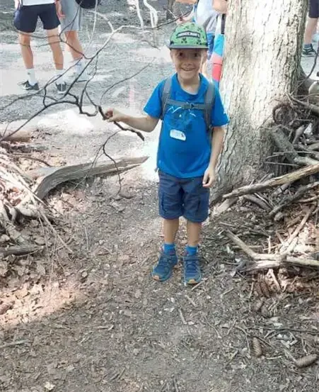 Ein kleiner Junge in einem blauen Shirt und einem grünen Hut hält einen Stock auf einem Wanderweg, mit anderen dahinter.