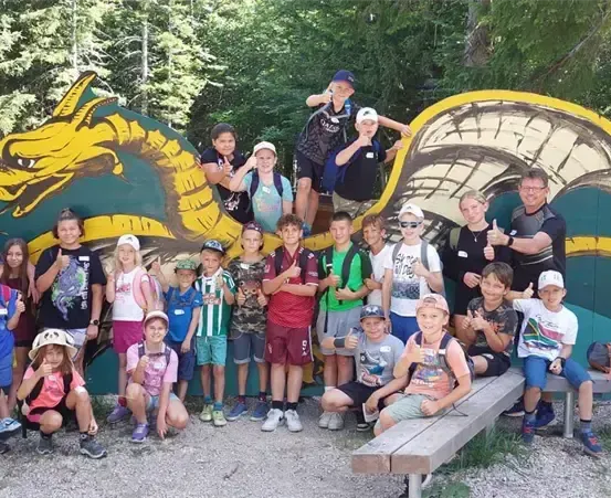 Eine Gruppe von Kindern und Erwachsenen steht vor einer Drachenmalerei im Wald, lächelt und posiert für ein Foto.