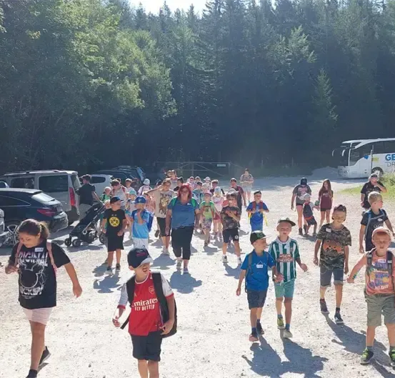 Eine Gruppe von Kindern und Erwachsenen geht in einer Reihe auf einem Erdweg, einige mit Rucksäcken, in einem Wald mit geparkten Autos und Bussen.