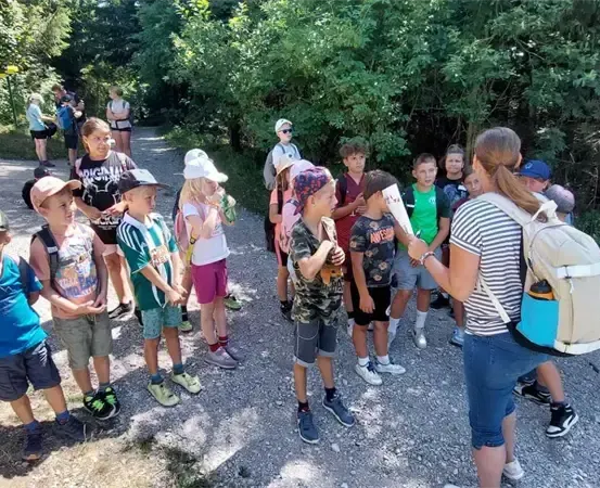 Eine Gruppe von Kindern und Erwachsenen steht auf einem Kiesweg, einige tragen Hüte und haben Rucksäcke dabei, in einem bewaldeten Gebiet. Eine Frau mit einem Rucksack spricht zu ihnen.