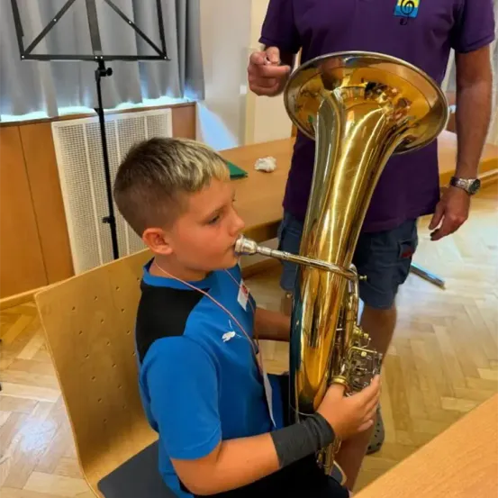 Ein junger Junge sitzt und spielt Tuba. Ein Erwachsener steht hinter ihm, möglicherweise um Anleitung zu geben. Die Umgebung umfasst einen Notenständer und einen Holztisch.