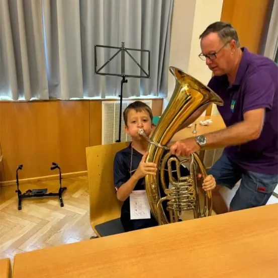 Ein junger Junge spielt Tuba, während ein Erwachsener ihm in einem Musikraum hilft. Dahinter steht ein Notenständer und ein Vorhang.