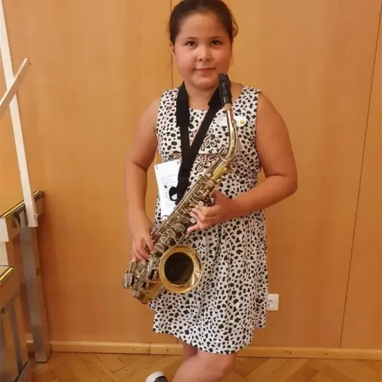 Ein junges Mädchen in einem gepunkteten Kleid posiert mit einem Saxophon, lächelt und steht in einem Raum mit Holzwänden und einem Holzboden.