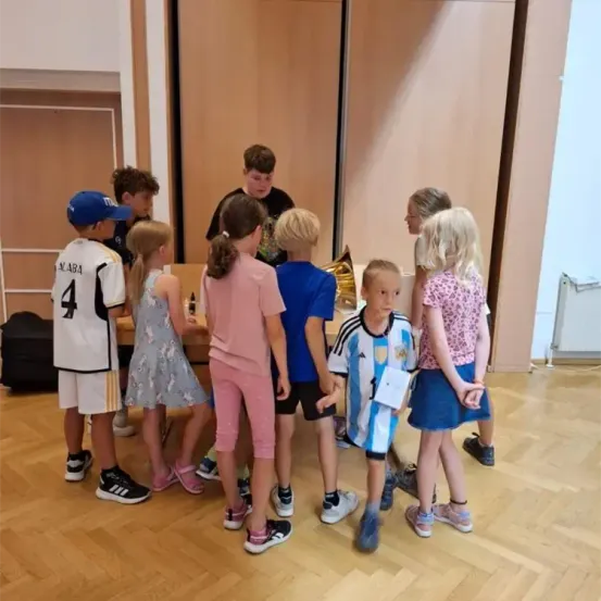 Mehrere Kinder stehen in einem Kreis um einen Tisch und nehmen wahrscheinlich an einer Veranstaltung teil. Ein Junge mit blauer Kappe und ein Mädchen in einem blauen Kleid sind anwesend. Ein Junge in einem Fußballtrikot hält ein Papier. Die Umgebung ist drinnen, mit einem Holzboden und einer Tür.