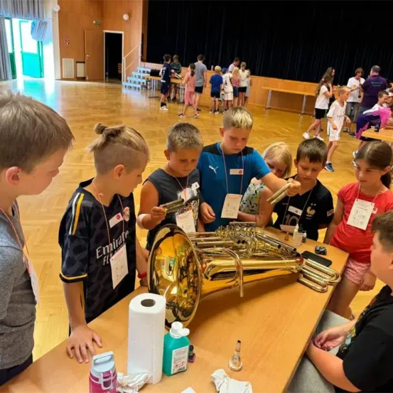 Kinder stehen um ein Tuba auf einem Tisch in einer Schulaula herum, tragen Namensschilder. Eine Gruppe von Kindern und Erwachsenen ist im Hintergrund, nahe einer Bühne und Tischen.