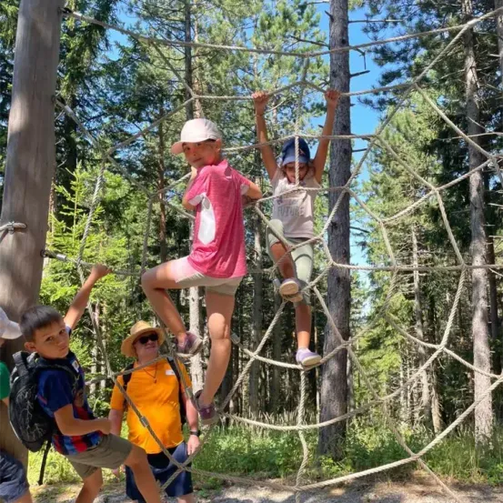 Eine Gruppe von Kindern klettert auf einer Seilleiter im Wald. Ein Kind trägt einen Hut und ein anderes eine Sonnenbrille.