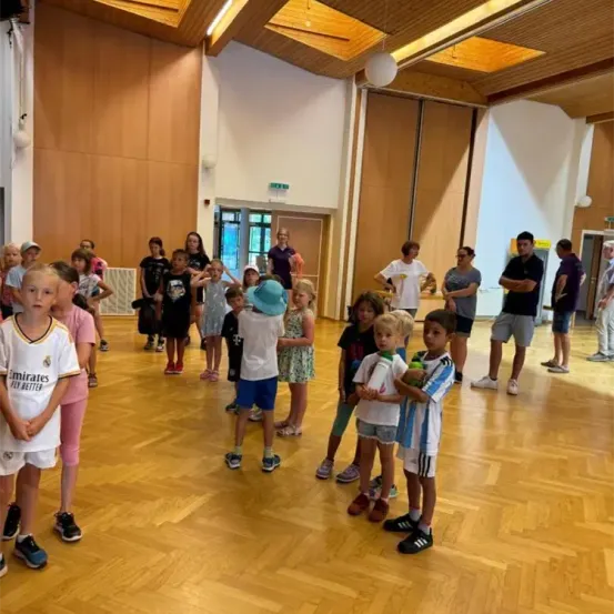 Eine Gruppe von Kindern und Erwachsenen versammelt sich in einem hölzernen Raum. Einige tragen Sporttrikots, und ein Kind hält einen Fußball. Hölzerne Wände und eine Decke mit hängenden Lampen schmücken den Raum.