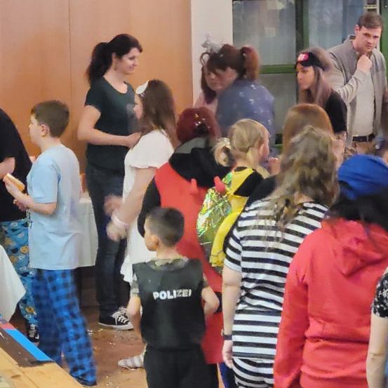 Eine Gruppe von Kindern und Erwachsenen versammelt, einige in Kostümen, einer mit einer Polizeiweste. Ein Junge hält Essen, andere unterhalten sich.