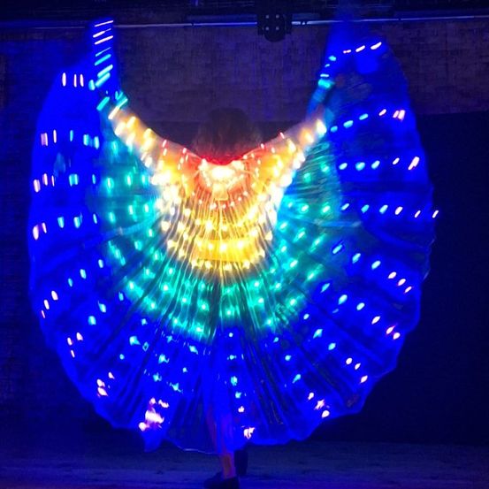Eine Person tanzt auf der Bühne mit einem großen, bunten LED-Ventilator. Der Ventilator leuchtet mit blauen, gelben und grünen Lichtern. Die Umgebung ist schwach beleuchtet.