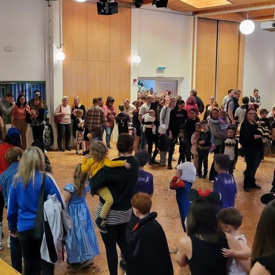 Eine große Gruppe von Menschen, darunter Erwachsene und Kinder, ist in einem Raum versammelt, wahrscheinlich für eine Halloween-Veranstaltung. Einige sind in Kostümen gekleidet.