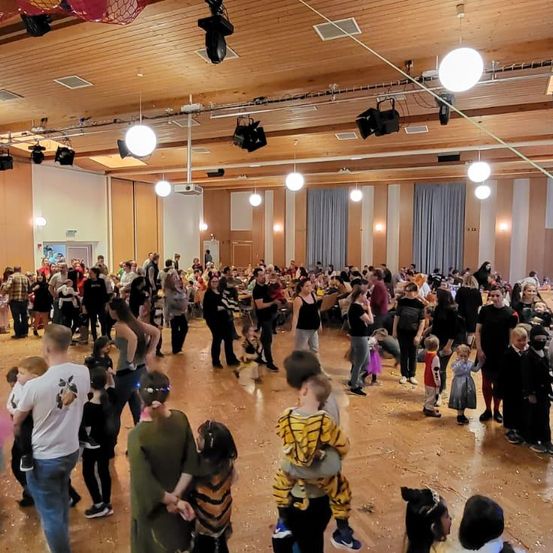 Ein überfüllter Saal mit vielen Menschen, einschließlich Erwachsener und Kinder, die an einer Aktivität teilnehmen. Der Raum ist hell beleuchtet und es gibt verschiedene dekorative Elemente.