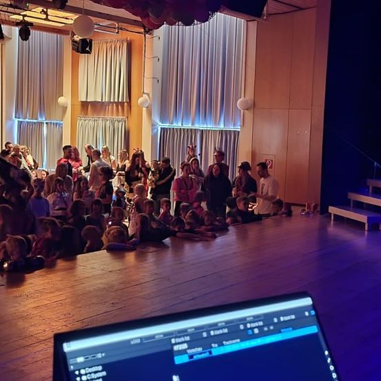 Eine große Indoor-Veranstaltung mit Menschen in Reihen, vermutlich für eine Aufführung. Die Bühne hat Lichter und ein Monitor zeigt etwas Text.
