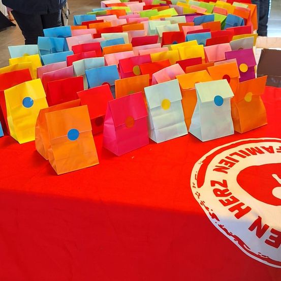 Ein mit rotem Tuch bedeckter Tisch ist mit vielen bunten Papiertüten mit kreisförmigen Designs gefüllt. Ein rotes Logo mit weißem Text befindet sich auf der rechten Seite des Bildes.