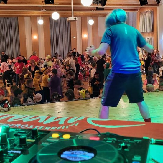 Ein Performer mit blauen Haaren steht auf der Bühne mit einer DJ-Anlage, während eine große Menschenmenge, einschließlich Kinder, zusieht. Die Bühne hat ein rotes und weißes Banner.