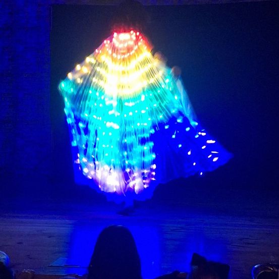 Eine leuchtende Lichtshow auf einem Stoffumhang beleuchtet eine Bühne mit blauen und mehrfarbigen Lichtern. Eine Person steht unter dem leuchtenden Umhang.