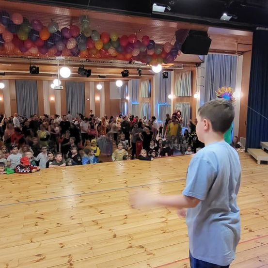 Ein Junge steht auf der Bühne in einem mit Luftballons dekorierten Saal vor einem Publikum aus Kindern in Kostümen. Der Saal hat Holzböden und blaue Vorhänge.