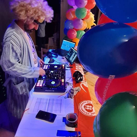 Ein Mann mit einer Perücke steht an einem DJ-Tisch mit Laptop, umgeben von bunten Luftballons.