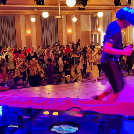 Eine Gruppe von Menschen versammelt sich in einem Saal mit einem DJ auf der Bühne, bunten Lichtern und Tischen. Einige Leute sitzen, andere stehen. Viele tragen Kostüme.