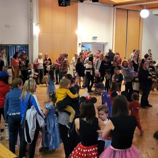 Viele Menschen sind drinnen versammelt, wahrscheinlich für eine Halloween-Veranstaltung. Einige sind in Kostümen, darunter ein Kind in einem Micky-Maus-Outfit.