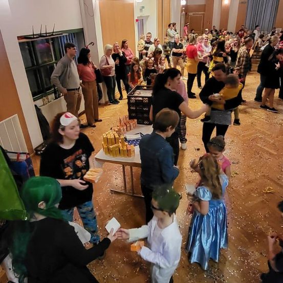 Eine große Menschenmenge, hauptsächlich Frauen und Kinder, ist drinnen versammelt. Einige halten Essen, während andere zuschauen. Der Raum ist mit Konfetti gefüllt.