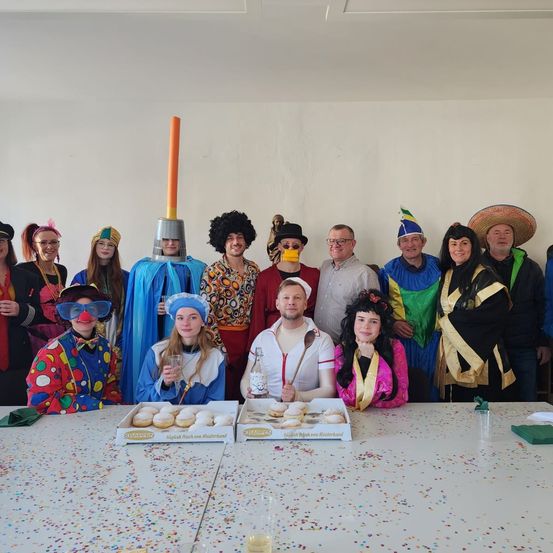 Gruppenfoto von Menschen in Kostümen, eine Person hält eine Flasche und eine andere einen Löffel, zwei Schachteln mit Donuts auf einem Tisch mit Konfetti.