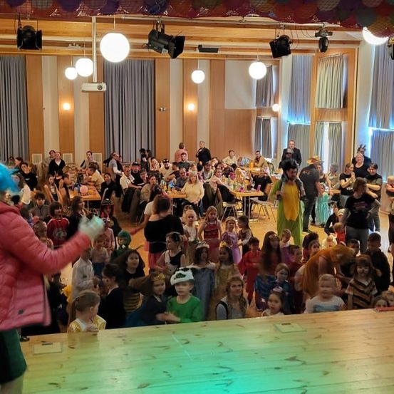 Eine große Menschenmenge aus Kindern und Erwachsenen versammelt sich in einem Saal. Eine Person in einer rosa Jacke führt die Menge an. Viele tragen Kostüme, einige haben Schminke im Gesicht. Tische mit Essen und Getränken sind im Saal aufgestellt.