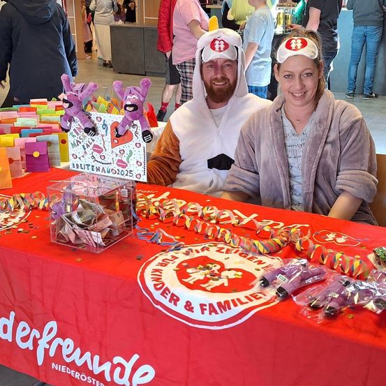 Zwei Menschen in Kostümen sitzen an einem Tisch mit Geschenken. Der Tisch hat eine rote Tischdecke mit einem Logo. Hinter ihnen stehen und gehen Menschen herum.