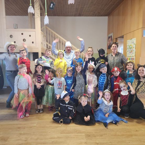 Eine Gruppe von Kindern und Erwachsenen in verschiedenen Kostümen posiert für ein Foto. Der Raum hat Holzwände und Böden, eine Treppe und ein hölzerne Spielhaus sind sichtbar.