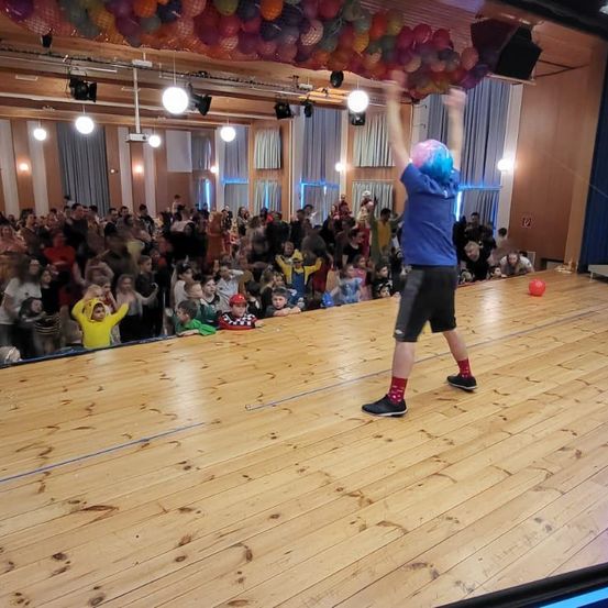 Eine Person mit blauen Haaren auf der Bühne, die Arme hebt, vor einem Publikum. Der Boden ist aus Holz, und die Decke hat Luftballons.