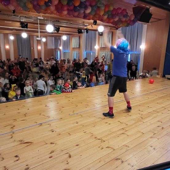 Eine Person mit blauen und rosa Haaren und einem blauen Shirt tritt auf einer Holzbühne auf, während ein Publikum zuschaut. Der Hintergrund ist mit Luftballons und Lichtern geschmückt.