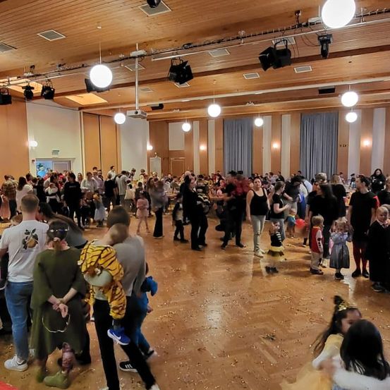 Eine große Gruppe von Menschen in einer Halle, viele in Kostümen, tanzen. Holzböden, etwas Konfetti, hängende Lampen.