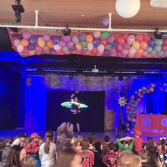Eine Darstellerin auf der Bühne mit einem leuchtenden Hula Hoop, umgeben von Luftballons und Bühnenlichtern. Darunter beobachtet ein Publikum von Kindern aufmerksam, einige tragen Mützen.